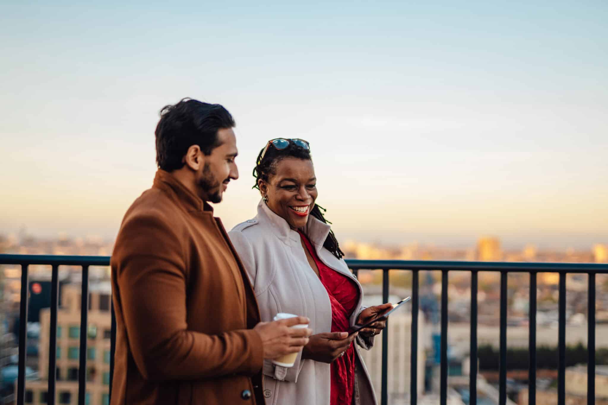 A man and woman holding coffee and a tablet on a rooftop balcony, planning their lifestyle goals through expert investment management.