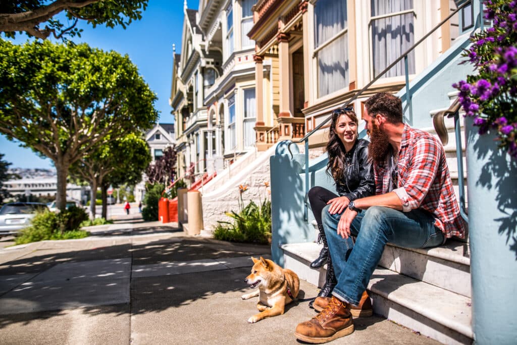 Lady and bearded man sitting on front stoop of a San Francisco house with a dog enjoying the sunshine.