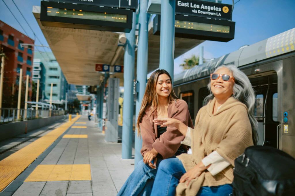 Two women, one younger and one older, are sitting and laughing together at an outdoor train platform.